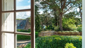 A view through a window of a garden with a large tree, a pathway, and a building with a sloped roof in the background. - Port Manech house 400 m from the beach, Pouldon cove 100 m away, small river view (Névez)