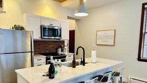 A modern kitchen with a stainless steel refrigerator, a microwave, a gas stove, and a sink. There are three white bar stools and a large pendant light above the island.