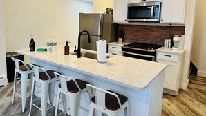 A modern kitchen with a central island, white bar stools, stainless steel appliances, and a brick backsplash.