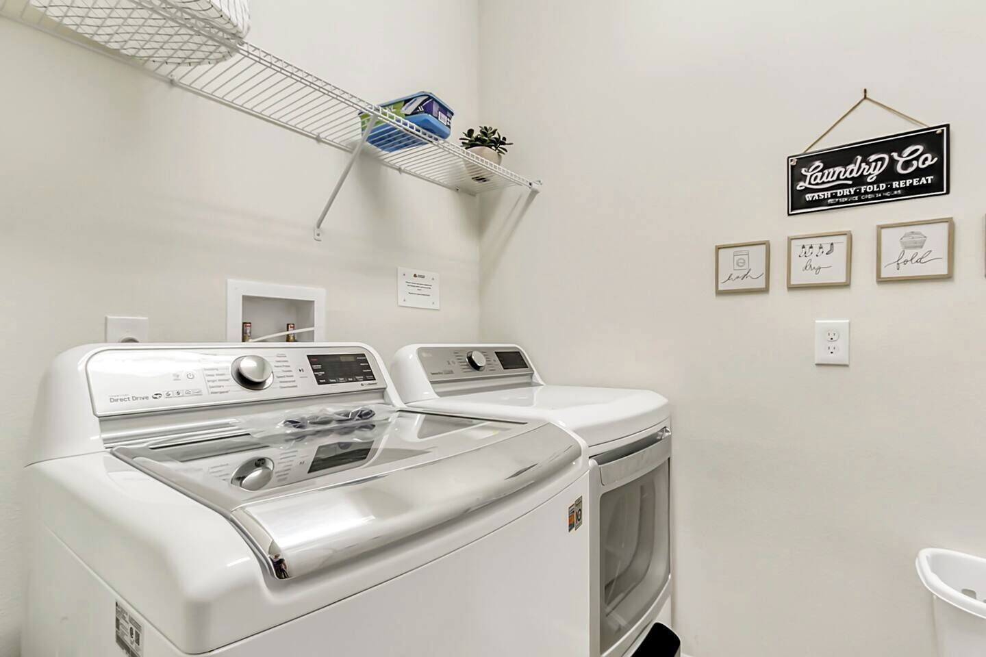 Uma lavanderia com duas máquinas de lavar e secar de abertura frontal, uma prateleira de metal com produtos de lavanderia e um aviso que diz "Laundry Co Wash Dry Fold Repeat".