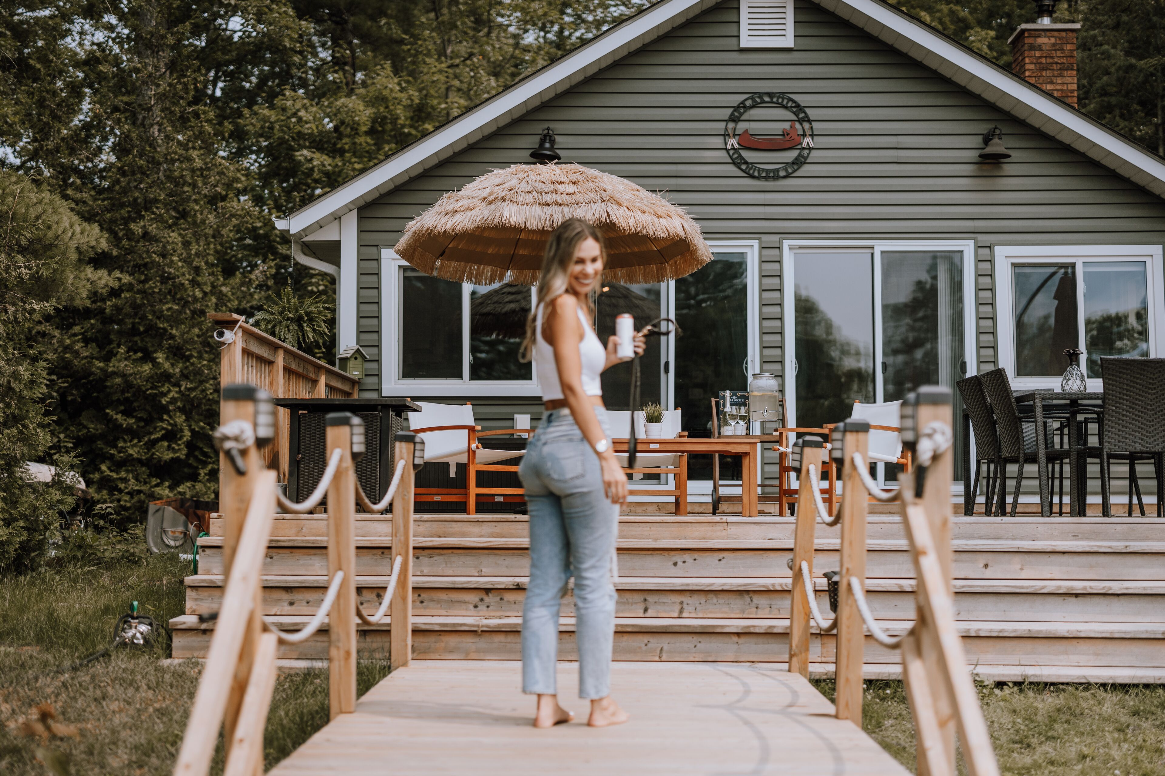 Une femme se tient sur une terrasse en bois devant une maison surmontée d’un parasol en chaume.