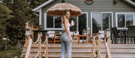 Une femme se tient sur une terrasse en bois devant une maison surmontée d’un parasol en chaume.