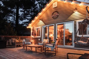 A wooden deck with outdoor seating, a hanging chair, and a well-lit interior visible through large windows. - Luxury Waterfront Cottage with Hot Tub in Downtown Washago, Ontario (Washago)
