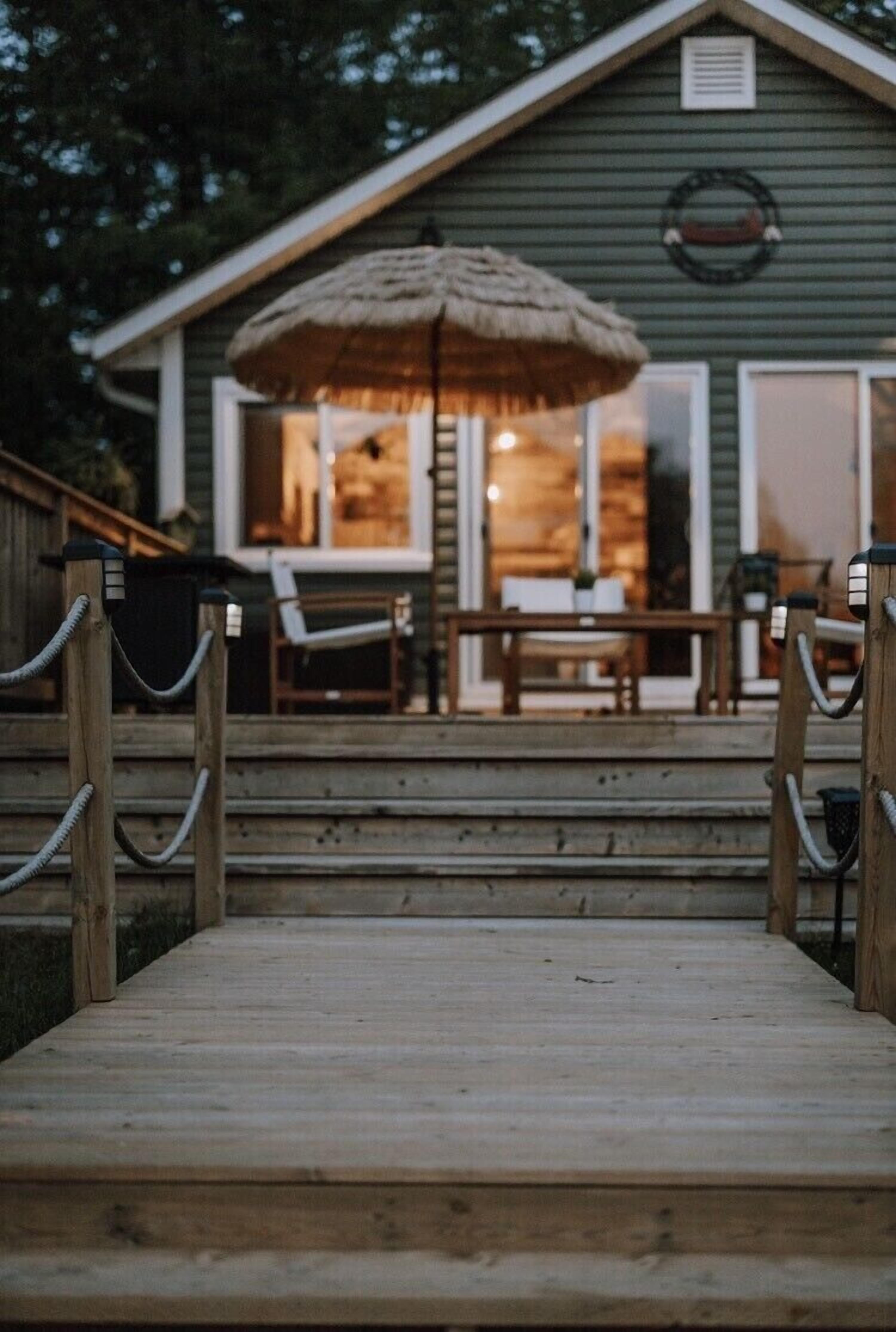 A wooden deck leading to a house with a thatched umbrella, outdoor furniture, and a visible window.