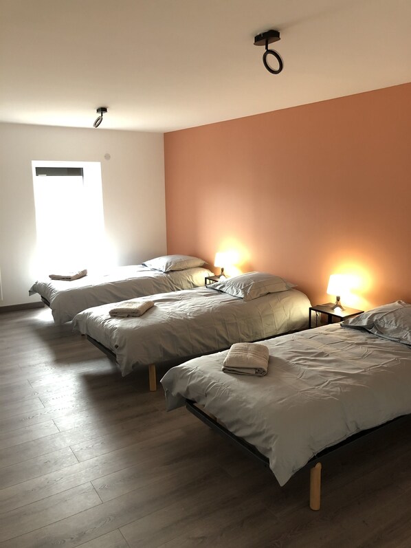 Une pièce avec trois lits alignés contre un mur de couleur pêche, un plancher en bois et une fenêtre laissant entrer la lumière naturelle.
