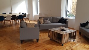 A modern living room with a grey sofa, a wooden coffee table, and a dining area with a white table and blue chairs. - Gîte Aux Portes du Sancy, en Auvergne, Jusqu'à 15 Personnes (Montaigut-le-Blanc)