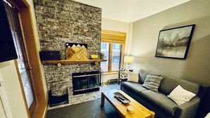 A living room with a stone fireplace, a sofa, a coffee table, and a window with blinds. - SKI-IN/OUT Charming studio condo steps to lift in the heart of Purgatory Resort! (Durango)