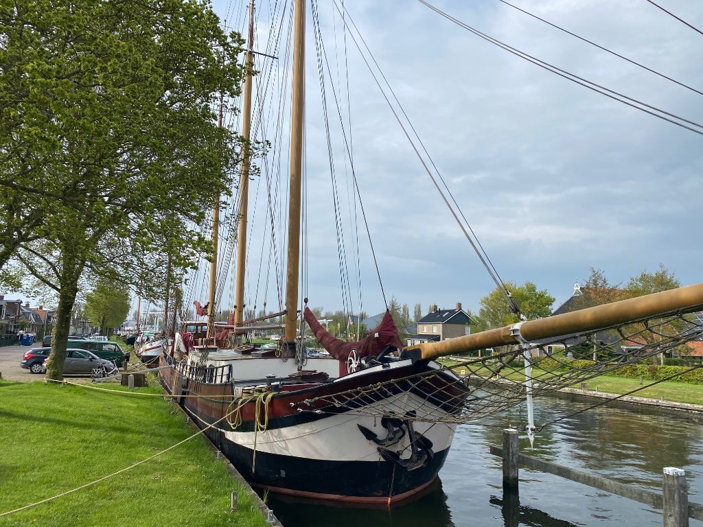 A historic sailing ship docked at a marina with a grassy area and parked cars nearby.
