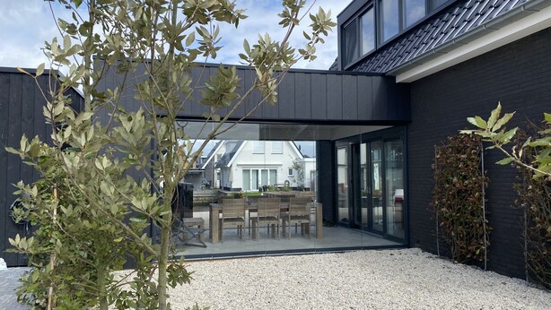 A modern outdoor patio area with a gravel floor, wooden furniture, and a view of a house through a glass wall. - Luxury vacation home including boat and heated veranda in Stavoren (Stavoren)