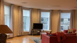 Een ruime woonkamer met een houten vloer, een rode bank, een televisie en grote ramen met gordijnen.