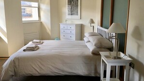 Een netjes opgemaakt bed met een witte deken en kussens, een nachtkastje met een lamp, een kleine witte ladekast en een ingelijste foto aan de muur.