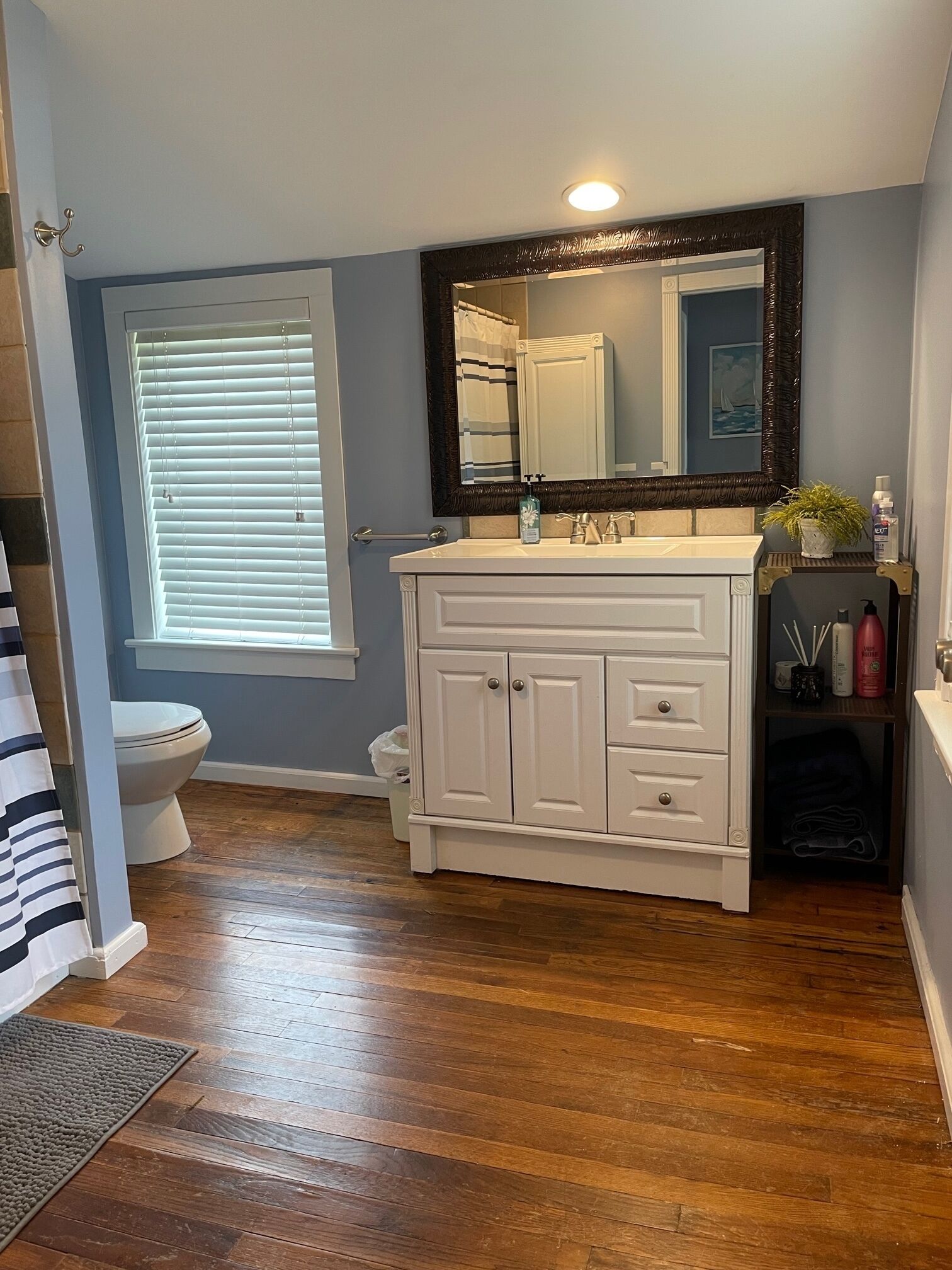 Une salle de bain avec un meuble-lavabo blanc, un grand miroir et une fenêtre avec des stores.