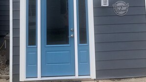 A building with a dark gray exterior, a blue door with a white frame, and a small window above the door. There is a welcome sign on the wall.