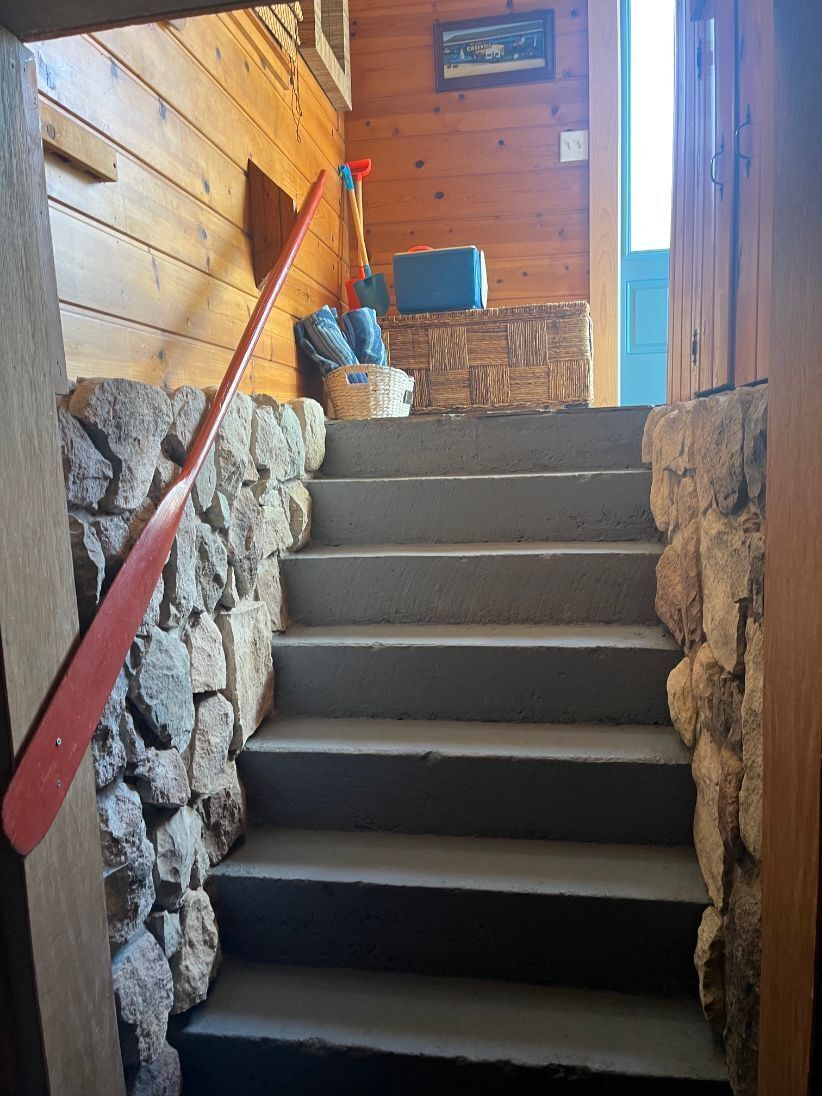 Un intérieur en bois avec un escalier en pierre, une rampe rouge et un panier contenant du matériel de nettoyage.
