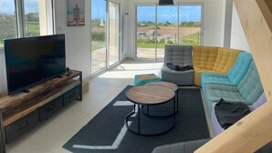 Un salon moderne avec un téléviseur à écran plat, un banc en bois, un fauteuil gris, un canapé jaune et bleu et une table basse ronde.
