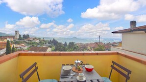 Un balcón con una mesa lista para dos personas, con vistas a un pueblo, un río y montañas a lo lejos.