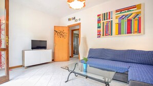 Una sala de estar con un sofá, una mesa de centro de vidrio, un televisor sobre un mueble y una llamativa obra de arte en la pared.