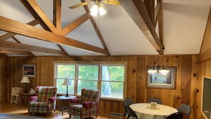 A cozy cabin interior with wooden walls, a dining table, chairs, a sofa, and large windows offering a view of trees. - Lovely spacious 6 BR home in St. Croix River valley (Taylors Falls)