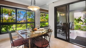 Un coin repas avec une table et des chaises en verre, un luminaire suspendu et une vue sur un jardin luxuriant et l’océan, le tout visible à travers de grandes fenêtres.
