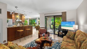 Un salon moderne avec un canapé, une table basse et un téléviseur. Il y a également un coin cuisine avec un bar et des chaises. La pièce est dotée de grandes fenêtres et offre une vue sur la verdure environnante.