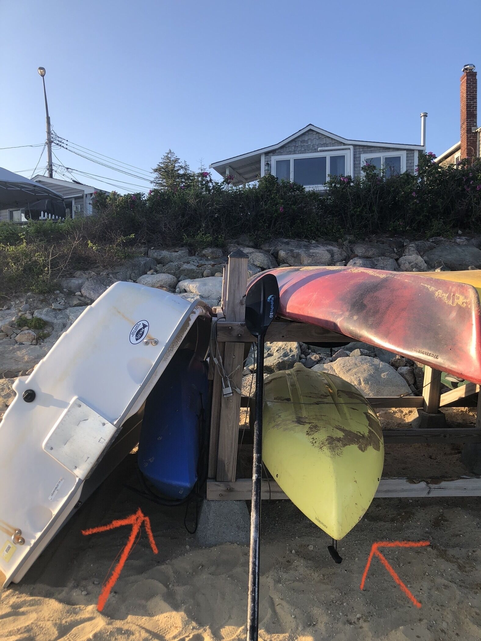 Kayaks alineados en un soporte de madera, con una playa arenosa y una casa al fondo.