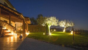 A well-lit outdoor area with a stone staircase leading to a wooden building, a lawn with a tree illuminated by lights, and a gravel path.