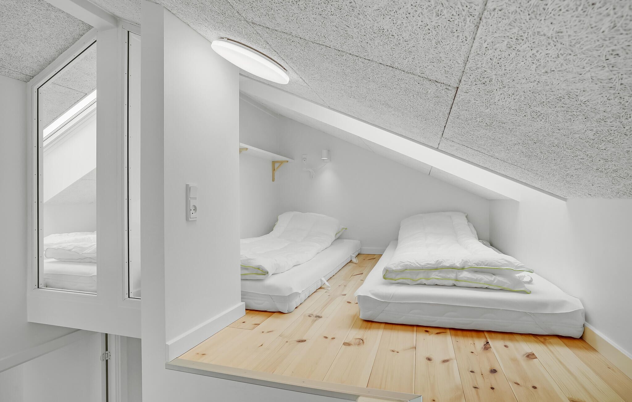 A compact, modern bedroom with a sloped ceiling, two single beds, a small bedside table, and a wall-mounted light fixture.