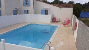 Uma piscina retangular com cerca branca, um flutuador salva-vidas vermelho à direita e uma fileira de cadeiras vermelhas ao longo da borda.