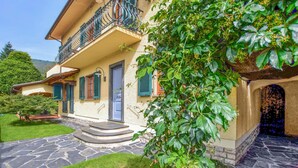 Ein zweistöckiges Haus mit einer Steintreppe, die zum Eingang führt, einem Balkon mit Metallgeländern und einem Garten mit einem Baum im Vordergrund.