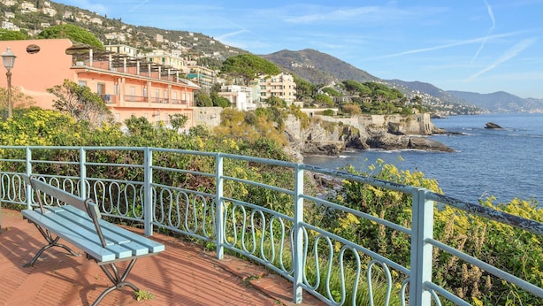 Uma vila costeira com edifícios numa encosta, um litoral rochoso e um banco em primeiro plano.
