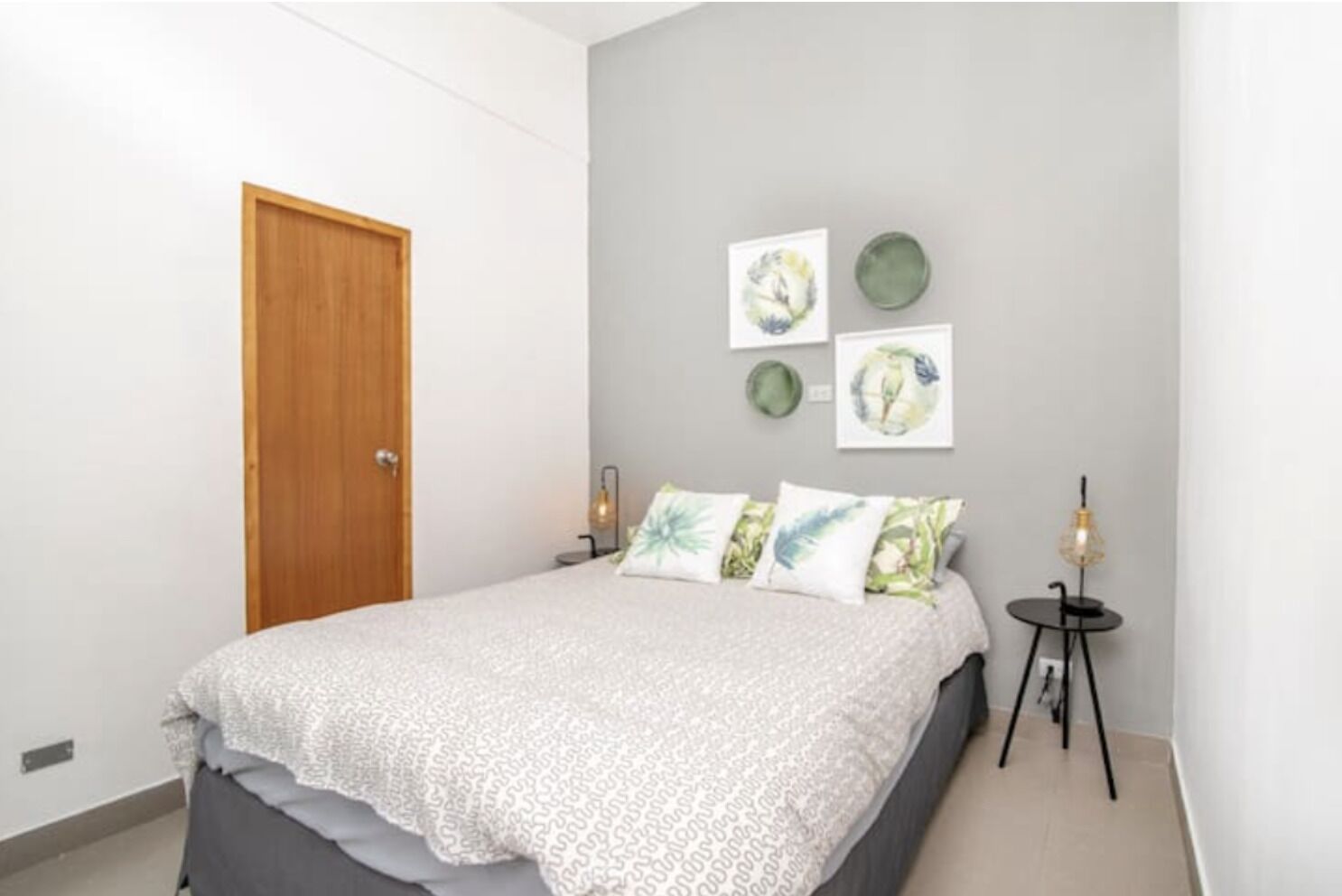 A bedroom with a bed, a wooden door, a wall with framed artwork, and a small table with a lamp.