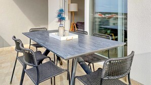 Ein rechteckiger Tisch im Freien mit dunkler Tischplatte, umgeben von sechs Korbstühlen mit Metallbeinen. Auf dem Tisch steht ein weißer Strauß mit blauen Verzierungen.
