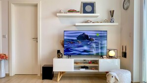 Ein Wohnzimmer mit Flachbildfernseher, einer weißen Unterhaltungsanlage, einer kleinen weißen Schaf-Dekoration und einem Fenster mit Blick ins Freie.