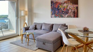 Ein modernes Wohnzimmer mit einem grauen Ecksofa, einem Glastisch und einem weißen Esstisch mit Stühlen. Ein großes Fenster bietet einen Ausblick nach draußen, eine Stehlampe und ein farbenfrohes abstraktes Gemälde schmücken die Wand.