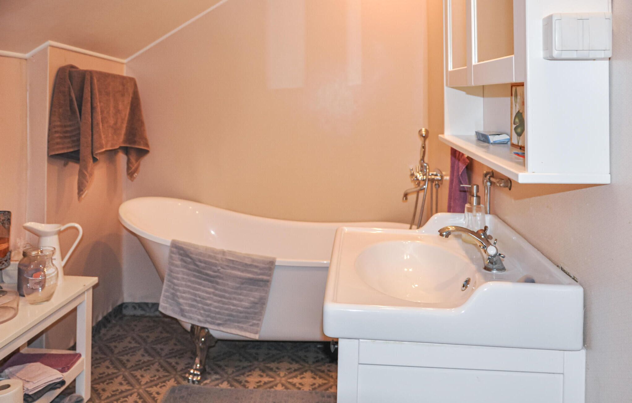 A bathroom with a white bathtub, a sink, and a mirror. There is a towel hanging on the wall and a shelf with toiletries.