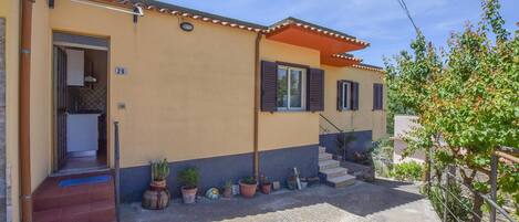Una casa de dos pisos con fachada color beige, el número '28' visible, un pequeño jardín delantero con plantas en macetas y una escalera que conduce a la entrada.
