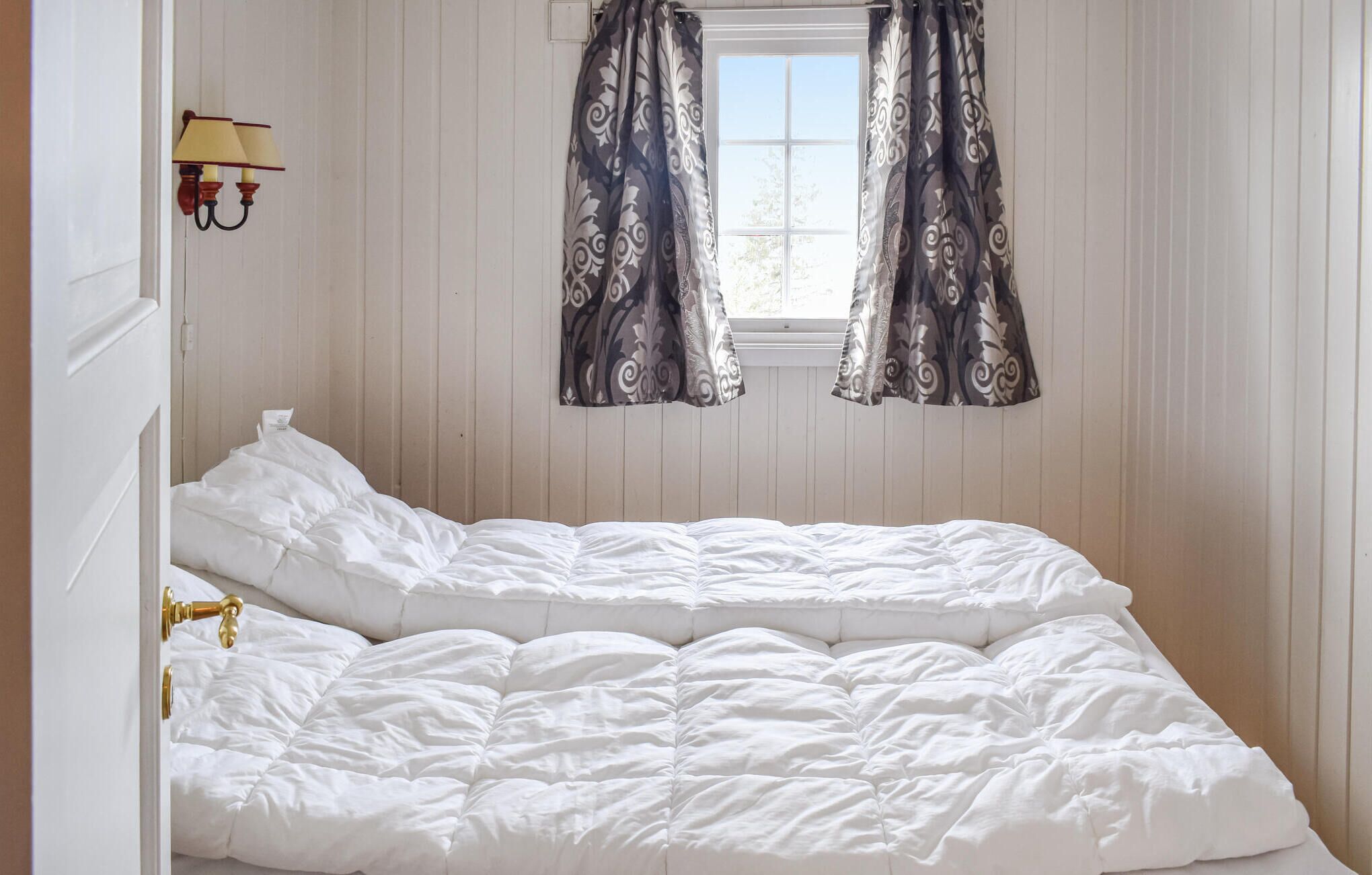 Une chambre à coucher avec un lit double, une fenêtre ornée de rideaux à motifs et un luminaire fixé au mur.