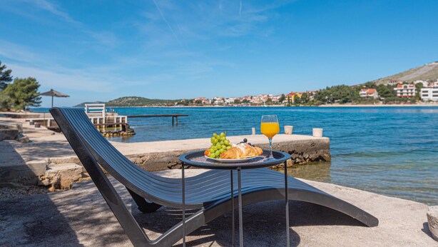 Uitzicht op zee met een gedekte tafel voor een maaltijd, inclusief druiven, een croissant en een glas sinaasappelsap, met zicht op een rustige zee en een kustplaats.