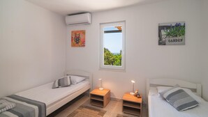 Quarto pequeno e bem iluminado com duas camas individuais, janela com vista para o verde, ar-condicionado fixado na parede e um letreiro com a palavra 'GARDEN'.