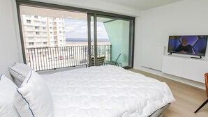 Ein Hotelzimmer mit einem großen Bett, einem Fernseher und einem Balkon mit Blick auf Gebäude und das Meer.