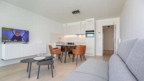 Ein modernes Wohnzimmer mit grauem Sofa, einer Essecke mit Tisch und Stühlen, einer Küche mit weißen Schränken und einem Fernseher an der Wand.