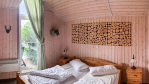 Ein Schlafzimmer einer Holzhütte mit Bett, Nachttisch, Lampe, einer Wanddekoration aus Baumstämmen und einem Fenster mit Vorhang.