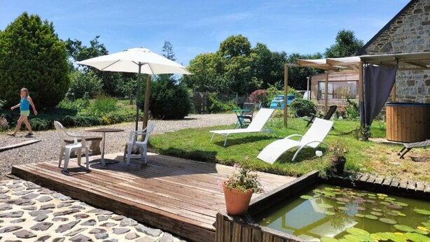 Una terraza de madera con sillones de descanso y una mesa, un pequeño estanque con nenúfares, y un edificio de piedra al fondo.