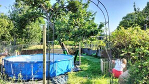 Un patio trasero con piscina azul, un parque infantil de madera y un área cercada con vegetación.