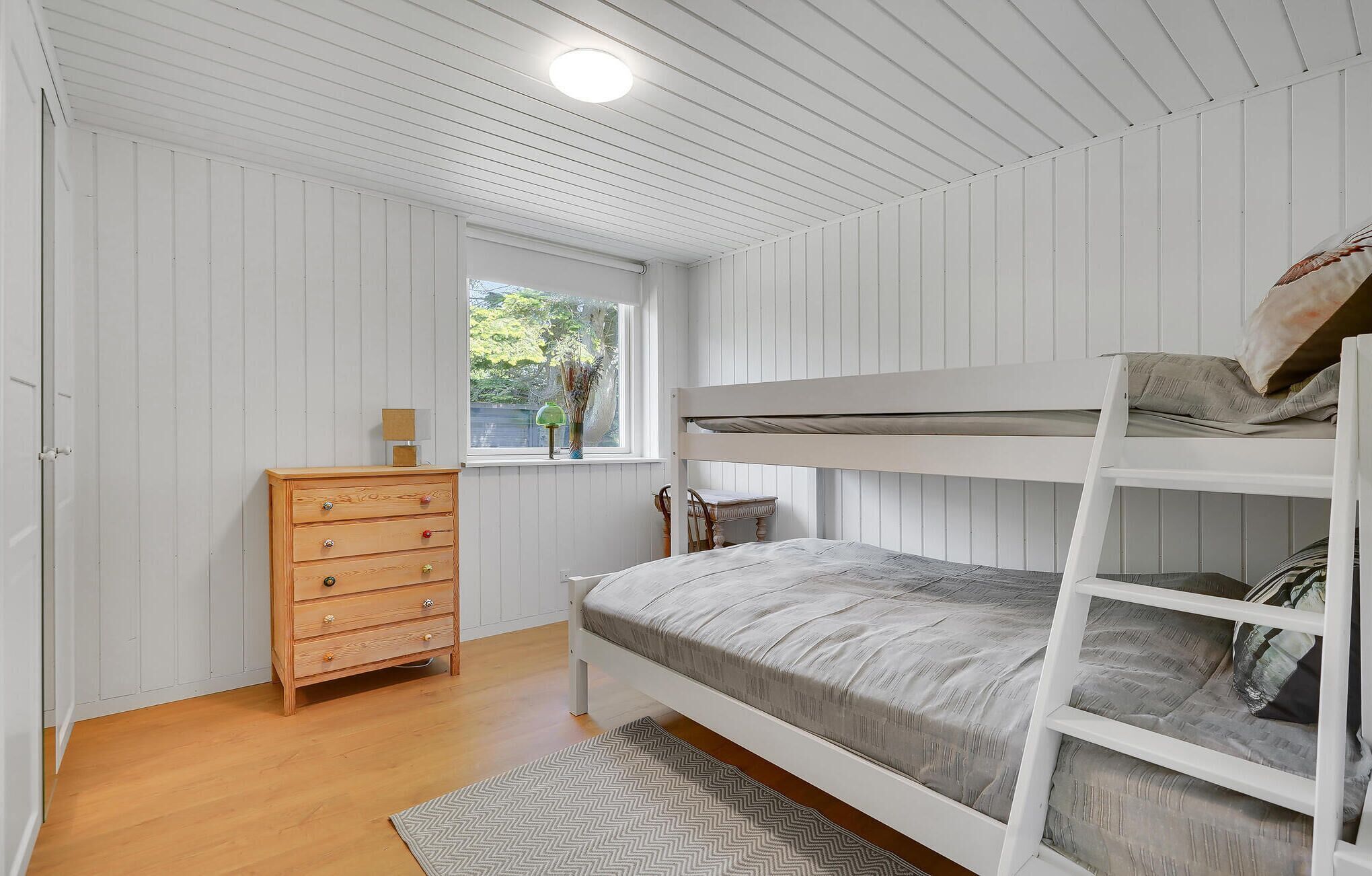 A bunk bed room with a wooden dresser, a window with a view of greenery, and a patterned rug.
