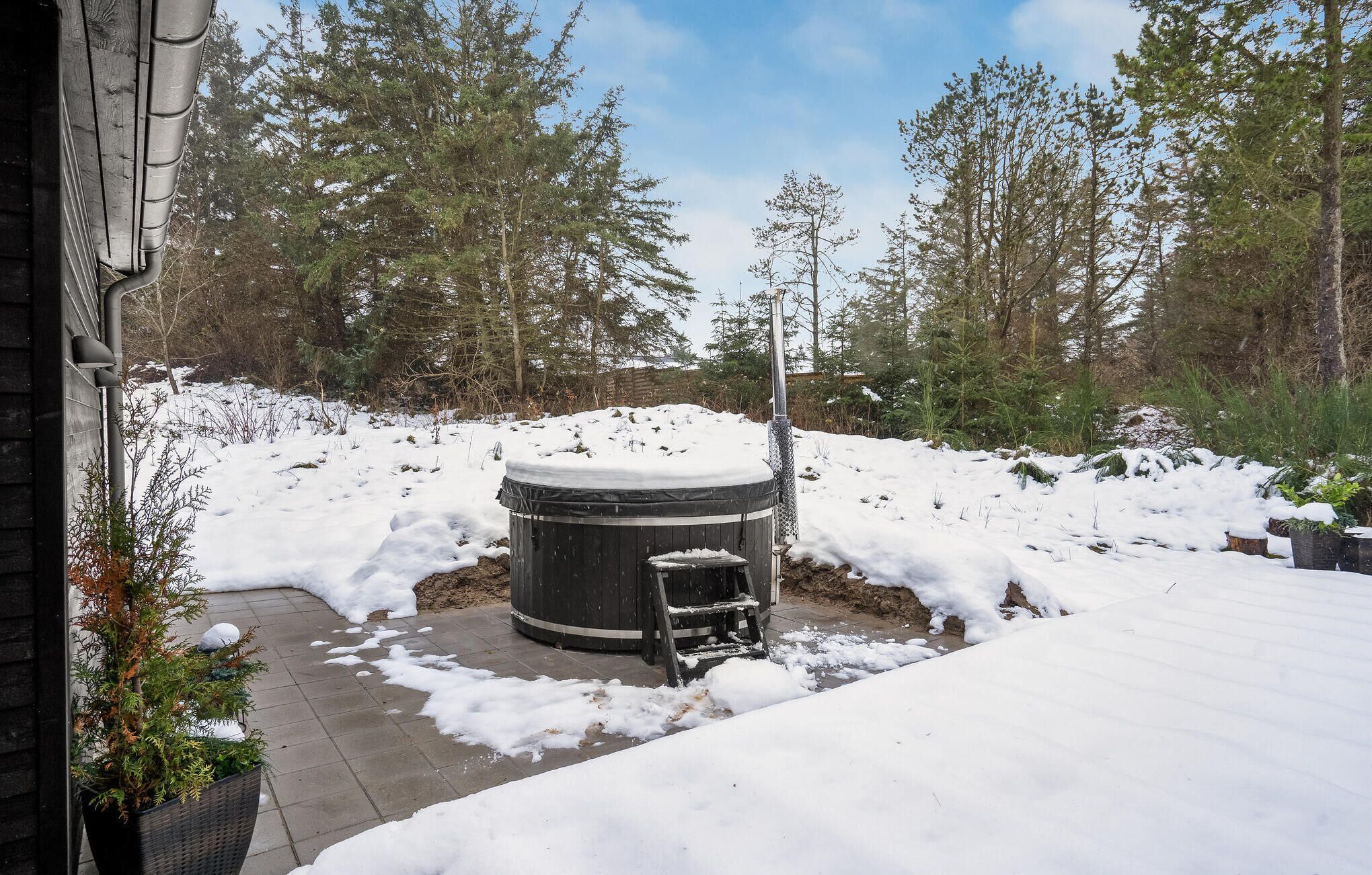 Un espace extérieur enneigé avec un jacuzzi, entouré d’arbres et d’une clôture en bois.