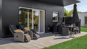 Een modern terras buiten met wicker meubilair, een glazen deur naar een tuin en een pergola met een parasol.