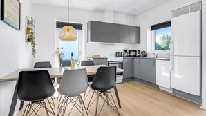 Een moderne keuken met grijze kastjes, een witte koelkast en een eethoek met een houten tafel en zwarte stoelen.