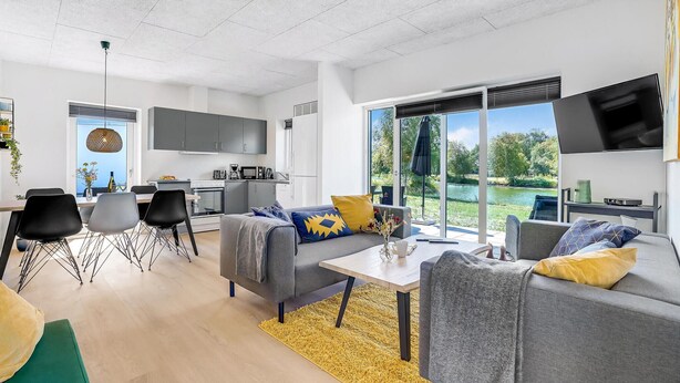 Een moderne woonkamer met een grijze bank, gele en blauwe kussens, een eethoek met een tafel en stoelen, en een keuken op de achtergrond.