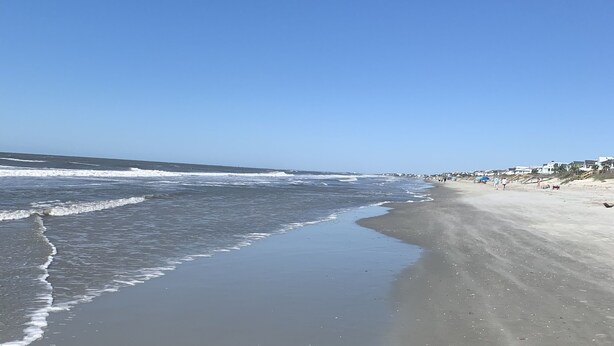 A wide sandy beach with gentle waves, a clear blue sky, and a distant shoreline with buildings. - Private home in Mt. Pleasant, SC Vacation Rental 10 min drive to the beach. (Mount Pleasant)